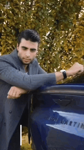 a man in a suit is leaning against a blue aston martin car .