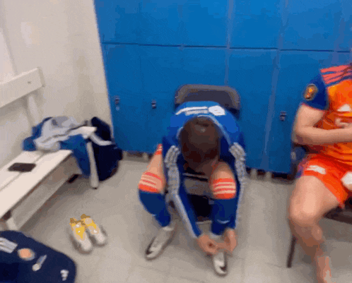 a man is tying his shoes in a locker room in front of blue lockers