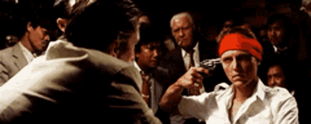 a man wearing a red headband is pointing a gun at another man 's head