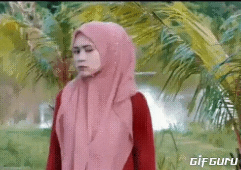a woman wearing a pink hijab and a red shirt is standing in front of palm trees .