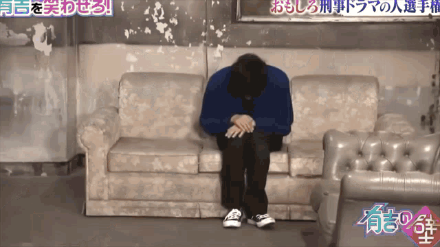 a man in a blue sweater sits on a couch with his head on his hands