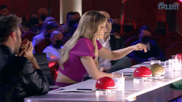 a woman in a purple top sits at a table in front of a talent show audience