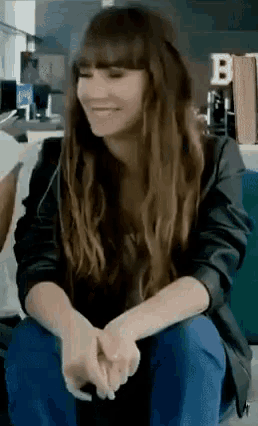 a woman with long brown hair is sitting on a couch with her hands folded and smiling .