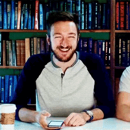 a man with a beard is smiling while holding a cell phone in front of a bookshelf
