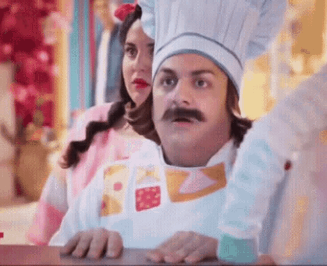 a man with a mustache is wearing a chef 's hat and apron while a woman looks on .
