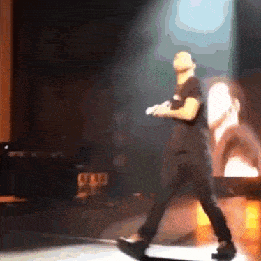 a man in a black shirt is walking on a stage with a blurred image of a man in a white shirt behind him