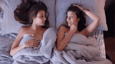 two women are laying in bed together under a blanket .