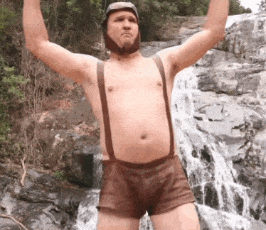 a shirtless man wearing suspenders and a helmet is standing in front of a waterfall
