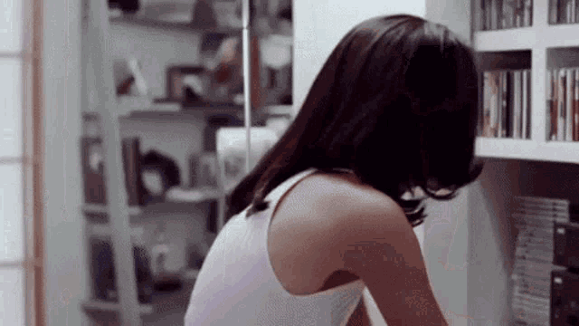 a woman in a white tank top is sitting in front of a bookshelf with cds on it .