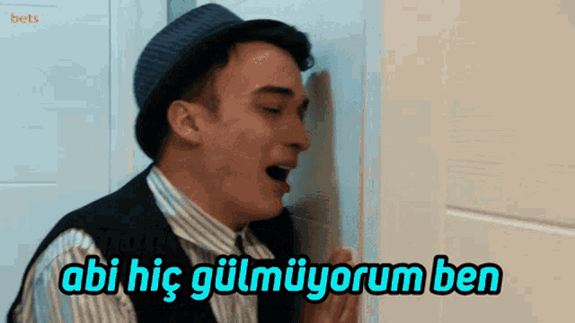 a man wearing a hat and a vest is talking into a door with the words abi hic gulmuyorum ben above him