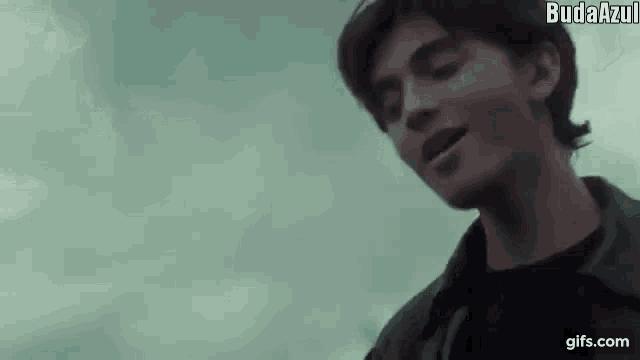 a young man is standing in front of a cloudy sky and singing into a microphone .