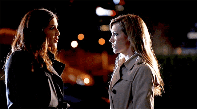 two women in trench coats are looking at each other at night