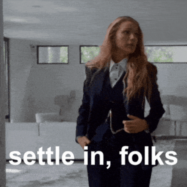 a woman in a suit is standing in a living room with the words " settle in folks " behind her