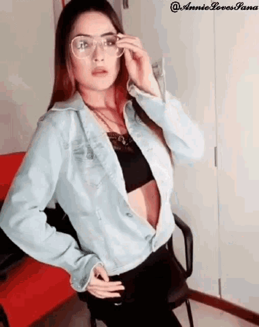 a woman wearing glasses and a denim jacket is sitting in a chair and adjusting her glasses .