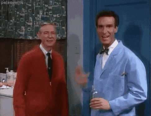 two men are waving at each other in a kitchen while one man is holding a glass of water .
