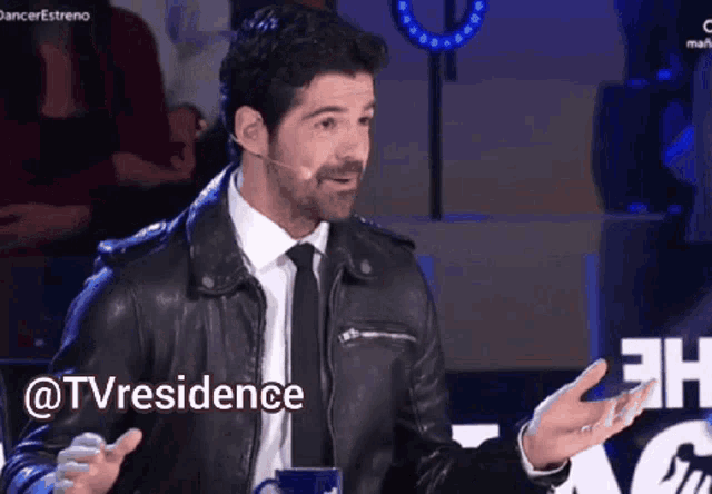 a man wearing a leather jacket and tie is talking into a microphone with the words @tvresidence written above him