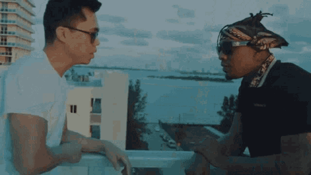 a man wearing sunglasses and a bandana talks to another man on a balcony overlooking a body of water