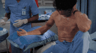 a shirtless man sits in a hospital bed with a nurse behind him with a name tag that says ' nurse '