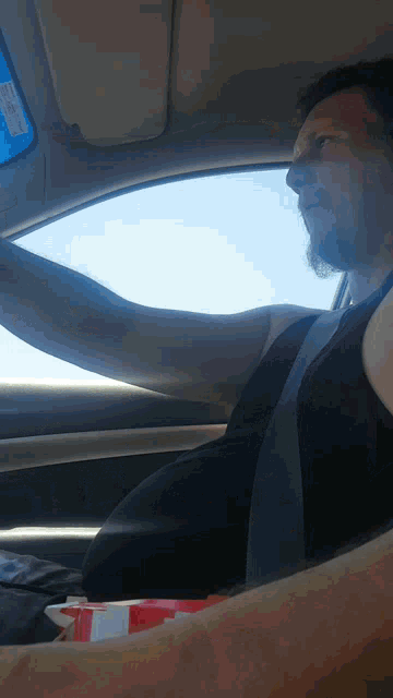a man in a black tank top is driving a car with a box of popcorn in his lap