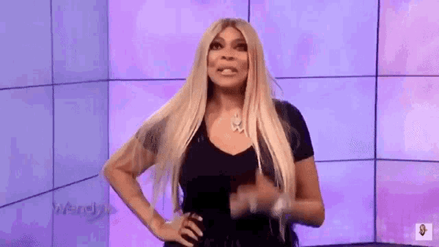 a woman with long blonde hair is standing in front of a purple wall and making a funny face .