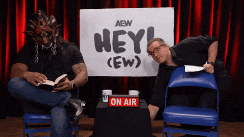 two men are sitting in front of a sign that says hey
