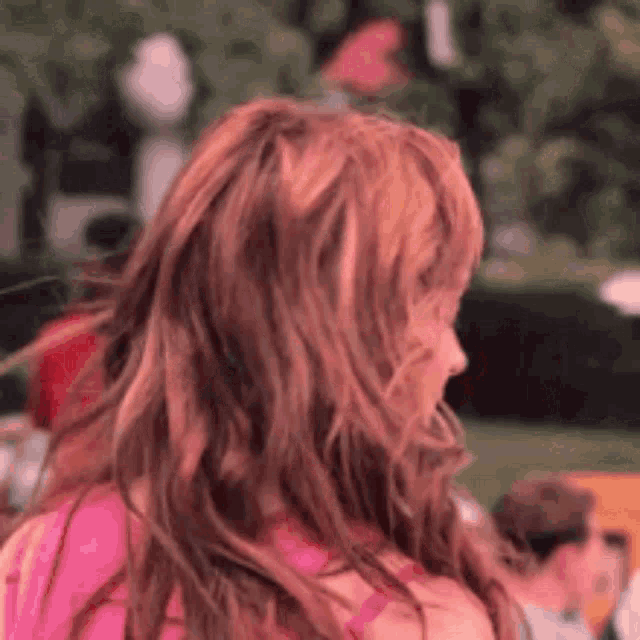 a woman with long red hair is standing in a park looking at the camera .