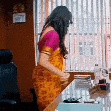 a woman in a yellow saree is standing in front of a desk with a calculator on it .