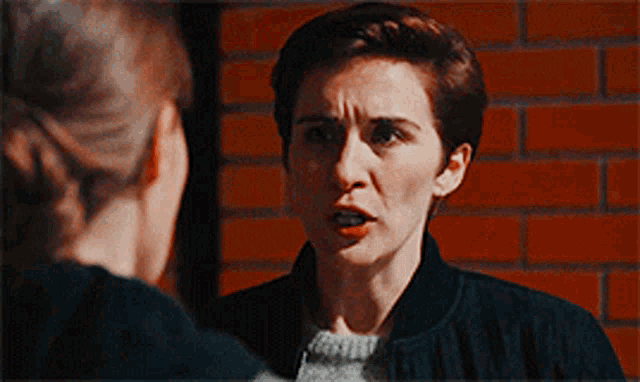 a woman in a black jacket is standing in front of a brick wall .
