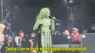 a woman in a green dress sings into a microphone with the words " today i am in drag of the biggest queen of all "