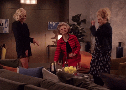three women are dancing in a living room with a bottle of wine on a table