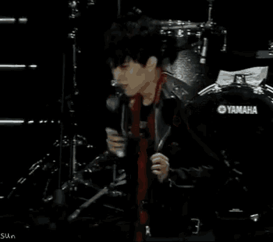a man in a red shirt is standing in front of a drum set and a microphone .
