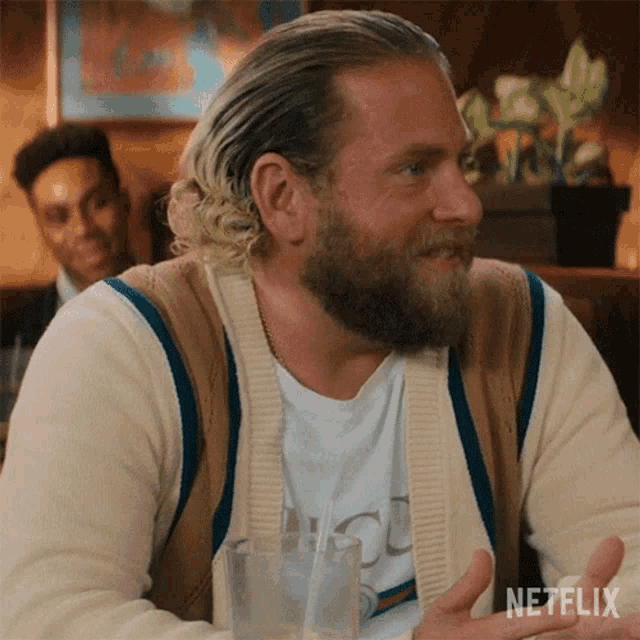 a man with a beard is sitting at a table with a glass of water and a netflix logo in the corner .
