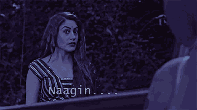 a woman in a striped top is talking to a man in a dark room and the word naagin is above her