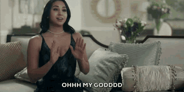 a woman in a black dress is sitting on a couch with her hands in the air and says ohh my godddd .