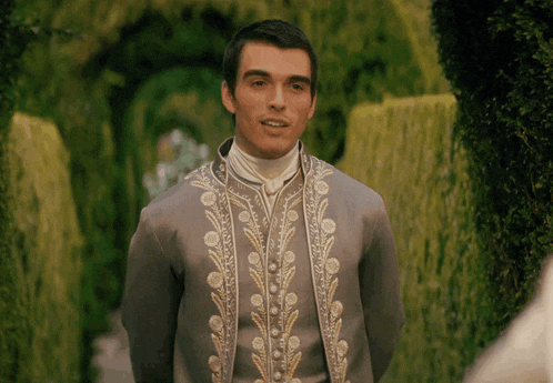 a man in a grey and gold embroidered suit stands in a garden