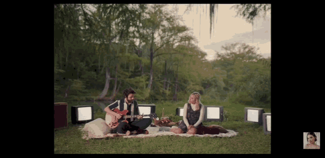 a man is playing a guitar while a woman sits on a picnic blanket
