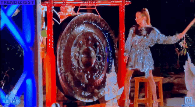 a woman in a silver dress is standing in front of a large gong that says trendisst on the screen