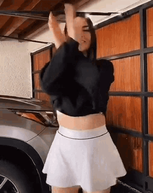 a woman in a black crop top and a white skirt is dancing in front of a car .