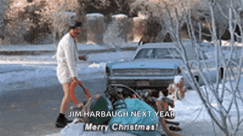 a man in a white shirt and shorts is standing in the snow next to a car .
