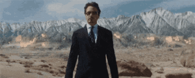 a man in a suit and tie is standing in a desert in front of a mountain range .
