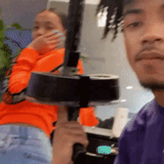 a man in a purple shirt is holding a black object in front of a woman in an orange sweater