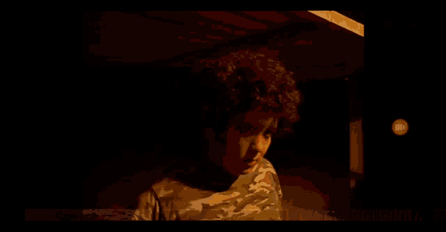 a young boy with curly hair is wearing a camo shirt and standing in a dark room .
