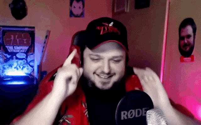 a man wearing a red shirt and a black hat is smiling in front of a rode microphone