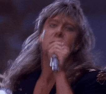 a man with long hair is singing into a microphone with his hands folded .