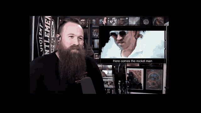 a man with a beard stands in front of a screen that says " here comes the rocket man "