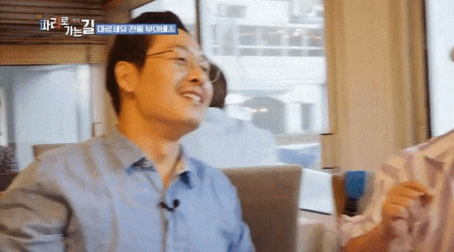 a man wearing glasses and a blue shirt is smiling in front of a window with a foreign language caption