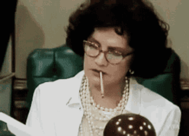 a woman wearing glasses and a pearl necklace is smoking a cigarette while reading a piece of paper .