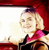 a woman is sitting in a red car looking out the window and smiling .