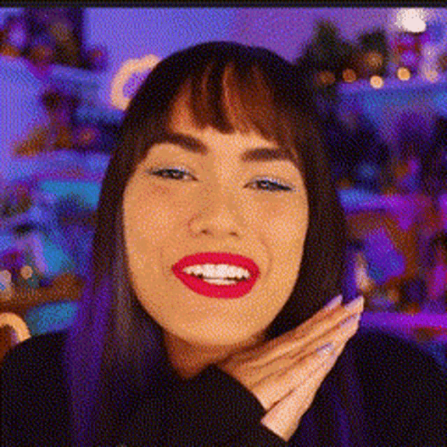 a woman with purple hair and red lipstick is smiling with her hands on her face .