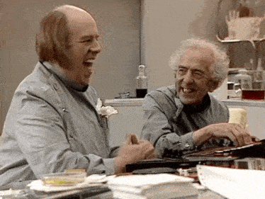 two men in lab coats are laughing in a lab while one is typing on a keyboard